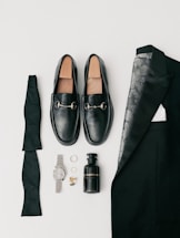 A collection of men's formal accessories neatly arranged includes black loafers with silver accents, a folded black bow tie, a black suit jacket with a patterned lining, a black bottle of cologne, a silver wristwatch, a pair of rings, and gold cufflinks.