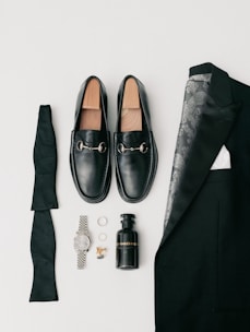 A collection of men's formal accessories neatly arranged includes black loafers with silver accents, a folded black bow tie, a black suit jacket with a patterned lining, a black bottle of cologne, a silver wristwatch, a pair of rings, and gold cufflinks.