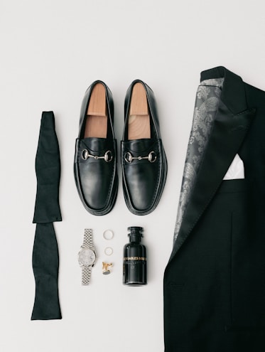 A collection of men's formal accessories neatly arranged includes black loafers with silver accents, a folded black bow tie, a black suit jacket with a patterned lining, a black bottle of cologne, a silver wristwatch, a pair of rings, and gold cufflinks.