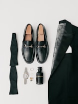 A collection of men's formal accessories neatly arranged includes black loafers with silver accents, a folded black bow tie, a black suit jacket with a patterned lining, a black bottle of cologne, a silver wristwatch, a pair of rings, and gold cufflinks.
