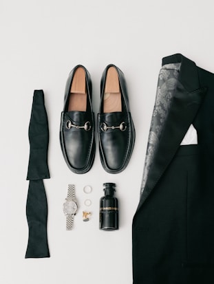 A collection of men's formal accessories neatly arranged includes black loafers with silver accents, a folded black bow tie, a black suit jacket with a patterned lining, a black bottle of cologne, a silver wristwatch, a pair of rings, and gold cufflinks.