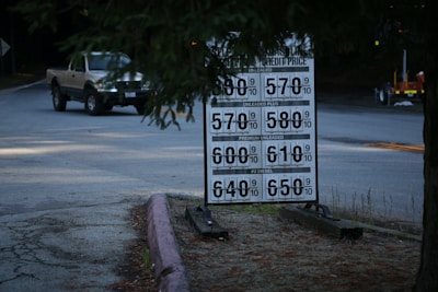 A roadside gas station price sign displays various fuel prices. To the left, a pickup truck is approaching on the road. The surroundings include trees and a dimly lit setting, indicating an evening or overcast atmosphere.