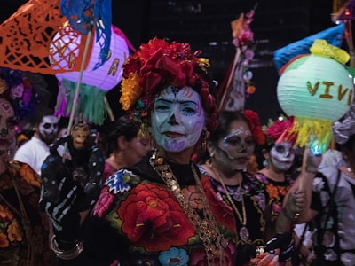 A group of people is dressed in traditional, colorful attire with intricate face paint resembling sugar skulls. They are holding vibrant paper lanterns and decorative crafts, appearing to celebrate a cultural event at night.