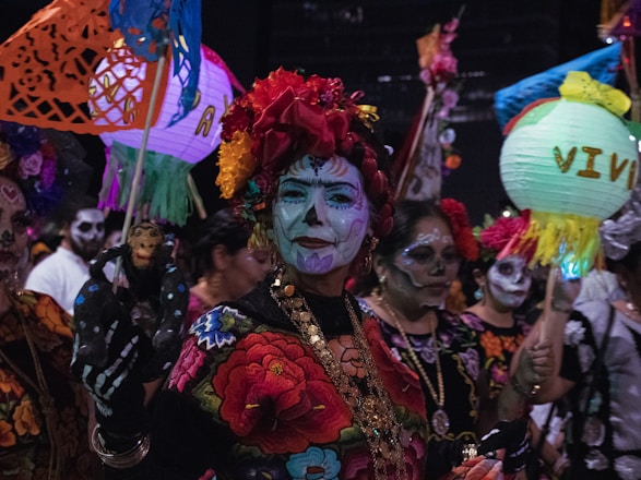 A group of people is dressed in traditional, colorful attire with intricate face paint resembling sugar skulls. They are holding vibrant paper lanterns and decorative crafts, appearing to celebrate a cultural event at night.