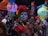 A group of people is dressed in traditional, colorful attire with intricate face paint resembling sugar skulls. They are holding vibrant paper lanterns and decorative crafts, appearing to celebrate a cultural event at night.
