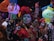 A group of people is dressed in traditional, colorful attire with intricate face paint resembling sugar skulls. They are holding vibrant paper lanterns and decorative crafts, appearing to celebrate a cultural event at night.