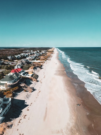 Aerial view of a beach with gentle waves breaking along the shore. Numerous houses and buildings are located close to the beach, extending parallel to the shoreline. The sky is clear and exhibits a vivid blue color.