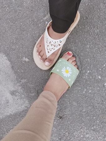 Two feet are visible, one wearing a white sandal with intricate cut-out designs and the other wearing a green slide with a daisy pattern and red painted toenails, standing on a rough asphalt surface.