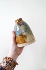 A hand is holding a handcrafted ceramic vase with a glazed finish. The vase features an irregular pattern with a mix of earthy tones, including brown, blue, and beige. The hand has a patterned sleeve that adds an artistic touch to the composition.