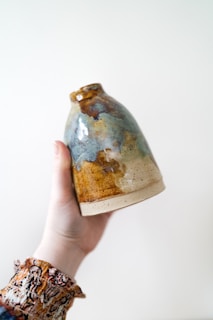 A hand is holding a handcrafted ceramic vase with a glazed finish. The vase features an irregular pattern with a mix of earthy tones, including brown, blue, and beige. The hand has a patterned sleeve that adds an artistic touch to the composition.