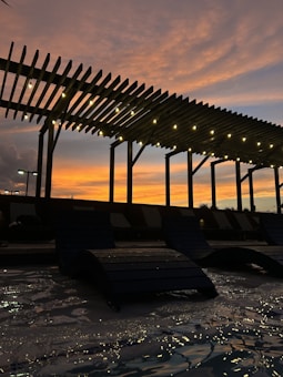 A pergola with lights is silhouetted against a vibrant orange and blue sunset. Beneath the structure, the water reflects the glowing lights and the colorful sky. Lounge chairs rest partially submerged in the water, creating a tranquil and relaxing atmosphere.