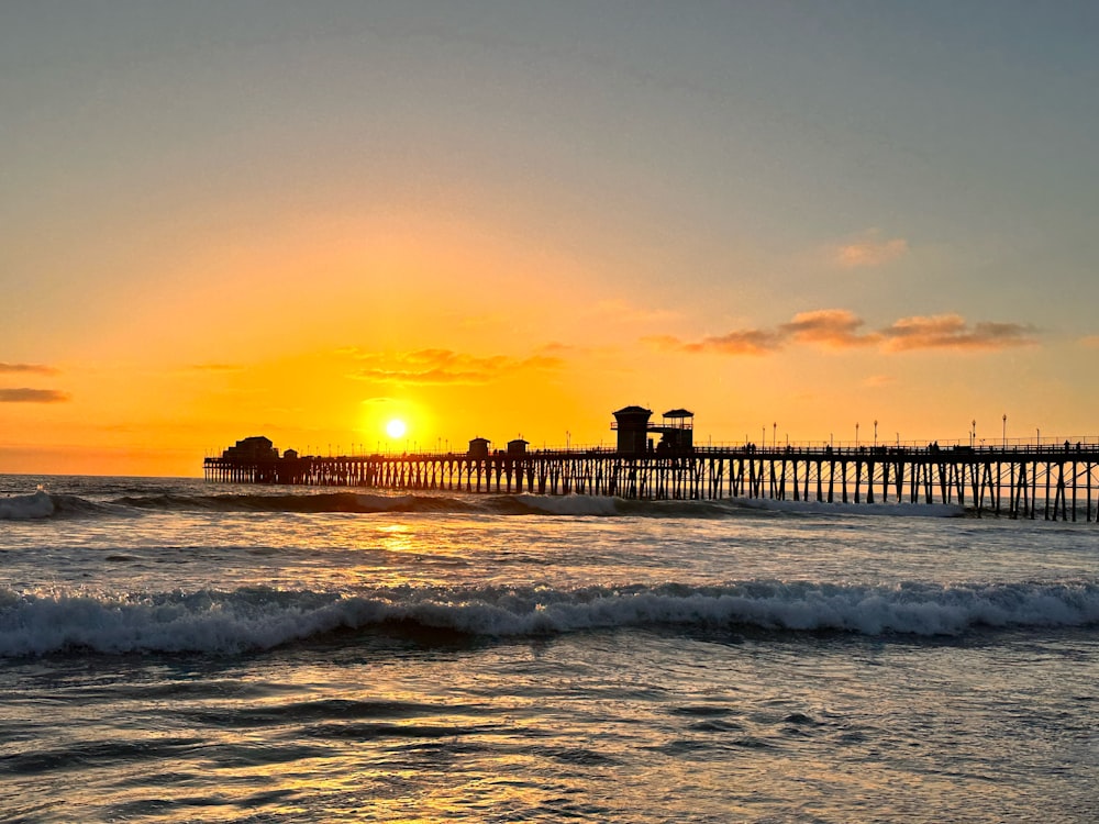 the sun is setting over the ocean with a pier in the background