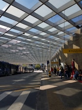 A modern bus station with a geometric pattern roof casting shadows on the ground. Several buses are lined up on the left side, while people are waiting near a platform with a yellow sign reading 'Plataforma.' The environment appears organized and functional, with clear pathways and a bus route.