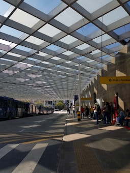 A modern bus station with a geometric pattern roof casting shadows on the ground. Several buses are lined up on the left side, while people are waiting near a platform with a yellow sign reading 'Plataforma.' The environment appears organized and functional, with clear pathways and a bus route.