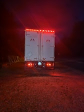 A large truck is parked on a gravel surface, illuminated by red taillights in a dimly lit environment. The vehicle has flags of Canada and the United States on its rear doors, with additional reflective markings and safety signs visible. The surrounding area is dark, hinting at nighttime or early evening.