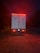 A large truck is parked on a gravel surface, illuminated by red taillights in a dimly lit environment. The vehicle has flags of Canada and the United States on its rear doors, with additional reflective markings and safety signs visible. The surrounding area is dark, hinting at nighttime or early evening.