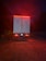 A large truck is parked on a gravel surface, illuminated by red taillights in a dimly lit environment. The vehicle has flags of Canada and the United States on its rear doors, with additional reflective markings and safety signs visible. The surrounding area is dark, hinting at nighttime or early evening.