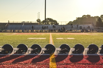 A line of black football helmets is neatly arranged on the edge of an American football field. In the background, a set of empty bleachers is visible with a few people scattered among the rows. The scene is bathed in warm, golden sunlight suggesting either early morning or late afternoon, with a clear sky and trees in the distance.