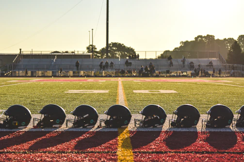 A row of clean, logo-free helmets lined up on a sports field bench under bright sunlight.
