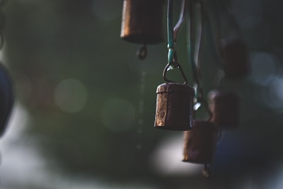 Hanging metal bells are suspended from cords, creating an artistic arrangement. The background is blurred, giving prominence to the bells, which have a rustic, weathered appearance. The ambiance is serene, emphasized by the soft light.