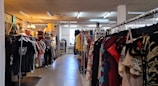 A clothing store interior featuring racks of diverse garments on both sides. The store includes various types of clothing, with organized sections and a mannequin dressed in casual attire. Warm lighting from hanging lamps creates a cozy atmosphere.