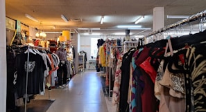 A clothing store interior featuring racks of diverse garments on both sides. The store includes various types of clothing, with organized sections and a mannequin dressed in casual attire. Warm lighting from hanging lamps creates a cozy atmosphere.