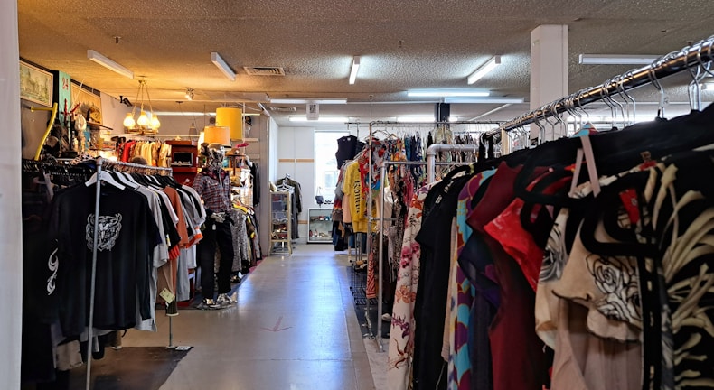 A clothing store interior featuring racks of diverse garments on both sides. The store includes various types of clothing, with organized sections and a mannequin dressed in casual attire. Warm lighting from hanging lamps creates a cozy atmosphere.