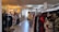 A clothing store interior featuring racks of diverse garments on both sides. The store includes various types of clothing, with organized sections and a mannequin dressed in casual attire. Warm lighting from hanging lamps creates a cozy atmosphere.