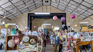 An indoor space filled with a variety of vintage items and collectibles displayed on shelves and racks. The ceiling is high and has a geometric pattern, adorned with hanging paper lanterns. A stage area named 'Broadway' is visible in the background, surrounded by an eclectic mix of objects including hats, vases, and framed pictures.