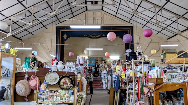 An indoor space filled with a variety of vintage items and collectibles displayed on shelves and racks. The ceiling is high and has a geometric pattern, adorned with hanging paper lanterns. A stage area named 'Broadway' is visible in the background, surrounded by an eclectic mix of objects including hats, vases, and framed pictures.