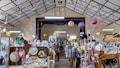 An indoor space filled with a variety of vintage items and collectibles displayed on shelves and racks. The ceiling is high and has a geometric pattern, adorned with hanging paper lanterns. A stage area named 'Broadway' is visible in the background, surrounded by an eclectic mix of objects including hats, vases, and framed pictures.