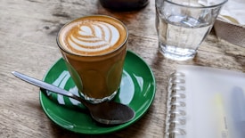 A glass of coffee with latte art on top is placed on a green saucer with a spoon resting on the side. Nearby, there is a clear glass of water and a spiral-bound notebook on a wooden table.