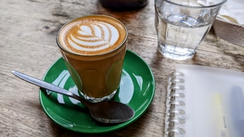 A glass of coffee with latte art on top is placed on a green saucer with a spoon resting on the side. Nearby, there is a clear glass of water and a spiral-bound notebook on a wooden table.
