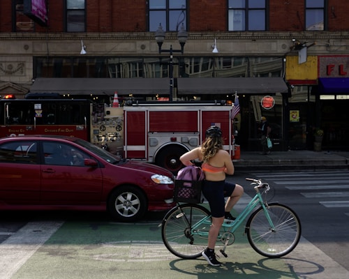 A person wearing a helmet and athletic clothing stands with a bicycle at a crosswalk. In front of them, a red car is stopped, and a fire truck with 'Chicago Fire Dept' is parked on the street. The background includes a brick building with large windows and various storefronts.