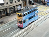 A vintage-style double-decker tram painted with ads is traveling on city streets, surrounded by modern buildings. The tram features a colorful mix of blue and orange with text and imagery promoting various products. The street includes multiple tracks and traffic markings, with a few pedestrians in the vicinity.