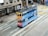 A vintage-style double-decker tram painted with ads is traveling on city streets, surrounded by modern buildings. The tram features a colorful mix of blue and orange with text and imagery promoting various products. The street includes multiple tracks and traffic markings, with a few pedestrians in the vicinity.