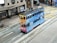A vintage-style double-decker tram painted with ads is traveling on city streets, surrounded by modern buildings. The tram features a colorful mix of blue and orange with text and imagery promoting various products. The street includes multiple tracks and traffic markings, with a few pedestrians in the vicinity.