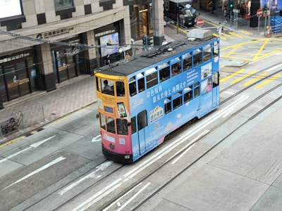 A vintage-style double-decker tram painted with ads is traveling on city streets, surrounded by modern buildings. The tram features a colorful mix of blue and orange with text and imagery promoting various products. The street includes multiple tracks and traffic markings, with a few pedestrians in the vicinity.