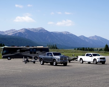 A scenic landscape with two trucks and a motorhome parked in a large open area. The trucks are towing trailers, and the backdrop features a range of mountains under a clear blue sky. Sparse trees and greenery surround the parking lot.