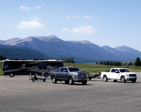 A scenic landscape with two trucks and a motorhome parked in a large open area. The trucks are towing trailers, and the backdrop features a range of mountains under a clear blue sky. Sparse trees and greenery surround the parking lot.