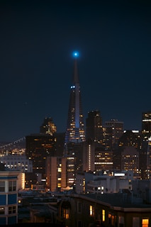 A nighttime cityscape depicting a skyline featuring a prominent pyramid-shaped skyscraper with a blue-lit tip. Surrounding the skyscraper are numerous other buildings with illuminated windows, contributing to an urban atmosphere. The scene conveys a modern, bustling city environment.
