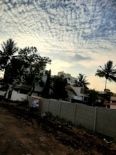 A residential area with a dirt road in the foreground and a concrete wall separating the road from several buildings. The sky is filled with scattered, fluffy clouds. Tall palm trees and other greenery are visible, adding a tropical feel to the scene. Overhead utility lines stretch across the sky.