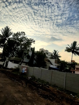 A residential area with a dirt road in the foreground and a concrete wall separating the road from several buildings. The sky is filled with scattered, fluffy clouds. Tall palm trees and other greenery are visible, adding a tropical feel to the scene. Overhead utility lines stretch across the sky.