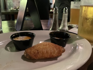 A close-up view of a plate with a single chicken wing placed in front of two small sauce cups. A glass of beer, salt and pepper shakers, and a dark table setting are visible in the background, suggesting a casual dining atmosphere.