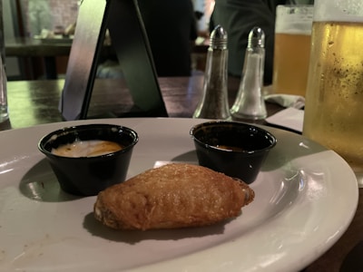 A close-up view of a plate with a single chicken wing placed in front of two small sauce cups. A glass of beer, salt and pepper shakers, and a dark table setting are visible in the background, suggesting a casual dining atmosphere.