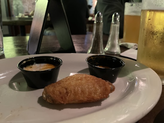 A close-up view of a plate with a single chicken wing placed in front of two small sauce cups. A glass of beer, salt and pepper shakers, and a dark table setting are visible in the background, suggesting a casual dining atmosphere.