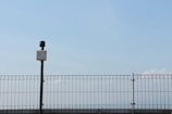 A metal fence under a clear blue sky with a surveillance camera positioned on top of a black pole. The pole and camera are centralized, with the fence extending across the width of the image. The sky is vast and mostly clear with a few faint clouds visible on the right side.