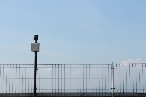 A metal fence under a clear blue sky with a surveillance camera positioned on top of a black pole. The pole and camera are centralized, with the fence extending across the width of the image. The sky is vast and mostly clear with a few faint clouds visible on the right side.