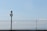 A metal fence under a clear blue sky with a surveillance camera positioned on top of a black pole. The pole and camera are centralized, with the fence extending across the width of the image. The sky is vast and mostly clear with a few faint clouds visible on the right side.