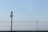 A metal fence under a clear blue sky with a surveillance camera positioned on top of a black pole. The pole and camera are centralized, with the fence extending across the width of the image. The sky is vast and mostly clear with a few faint clouds visible on the right side.